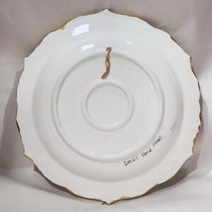 Puede incluir: Parte posterior de un plato de cer&aacute;mica blanco con borde festoneado y ribete dorado. Se adjunta una cuerda marr&oacute;n para colgarlo. Las palabras "Special Hand Made" est&aacute;n escritas en el plato.