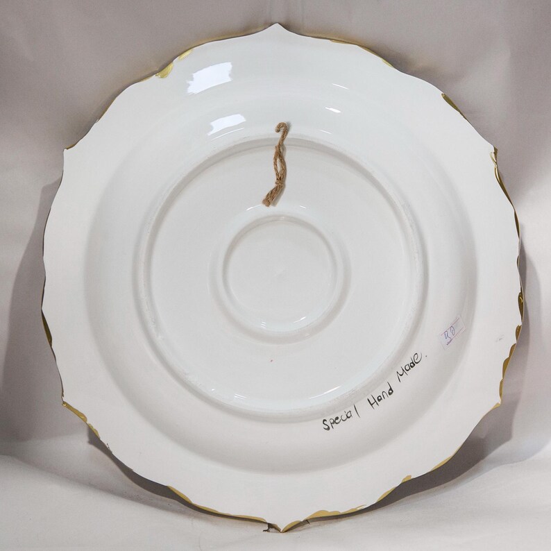 Puede incluir: Parte posterior de un plato de cer&aacute;mica blanco con borde festoneado y ribete dorado. Se adjunta una cuerda marr&oacute;n para colgarlo. Las palabras "Special Hand Made" est&aacute;n escritas en el plato.