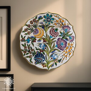 Puede incluir: Un plato decorativo de cer&aacute;mica con un dise&ntilde;o floral. El plato presenta un fondo blanco con flores coloridas en tonos de azul, morado, amarillo y rojo. El plato tiene un borde dorado y un borde festoneado. El plato est&aacute; montado en una pared beige.