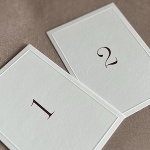 Peut inclure: Deux cartons rectangulaires blanc cassé avec un fin bord en relief. Les cartons sont numérotés 1 et 2 dans une police serif marron foncé. Les cartons sont présentés sur une surface aux tons neutres, suggérant un thème de mariage ou d'événement.
