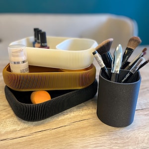 Könnte beinhalten: Eine Anordnung von Beauty-Organisatoren und Zubehör. Das Bild zeigt drei gerippte Tabletts in Creme, Gold und Schwarz, die Make-up und Hautpflegeprodukte enthalten. Ein schwarzer zylindrischer Halter enthält verschiedene Make-up-Pinsel. Ein orangefarbener Schwamm liegt im unteren Tablett.
