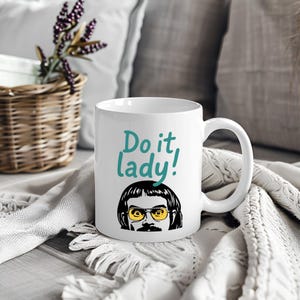 May include: White ceramic mug with the text "Do it, lady!" in teal script. Below the text is a cartoon illustration of a person with yellow-tinted glasses, a mustache, and dark hair. The mug has a curved handle.