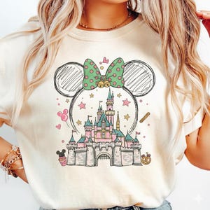 May include: Cream-colored t-shirt featuring a whimsical illustration of a castle, stars, and a Minnie Mouse-inspired bow with polka dots. The design includes a castle, stars, and a pretzel. The shirt has a crew neck.