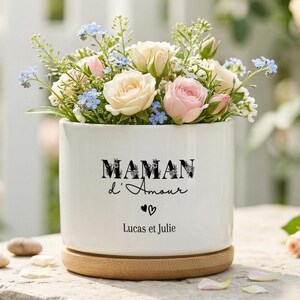 Könnte beinhalten: Ein weißer Keramiktopf mit Holzsockel, gefüllt mit einem Strauß aus rosa und cremefarbenen Rosen, blauen Vergissmeinnicht und Grünzeug. Der Topf trägt den Text "MAMAN d'Amour" und die Namen "Lucas et Julie".