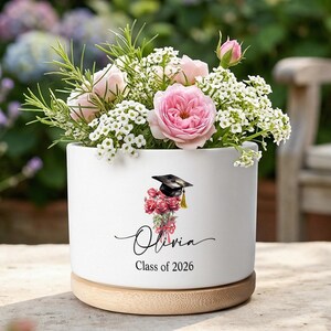 Könnte beinhalten: Ein weißer Keramiktopf mit Holzsockel, gefüllt mit rosa Rosen und weißen Blumen. Der Topf ist mit einer Abschlusskappe und dem Namen "Olivia" mit "Klasse 2026" darunter verziert. Der Topf befindet sich im Freien.
