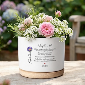 May include: A white ceramic planter with a wooden base, filled with pink roses, white flowers, and greenery. The planter has the text "Chapter 60" and a personalized name. The text also includes the year "EST. 1966".