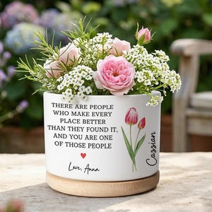 May include: A white ceramic flower pot with a wooden base, filled with pink roses and white flowers. The pot has a printed message and a tulip illustration. The text reads "There are people who make every place better than they found it, and you are one of those people."