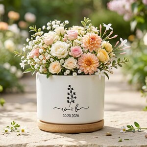 Può includere: Un vaso cilindrico in ceramica bianca con base in bambù, riempito con un bouquet di fiori rosa, pesca e bianchi. Il vaso presenta un disegno stampato in nero di un rametto, le lettere w + b e la data 20.10.2026 sulla parte anteriore.