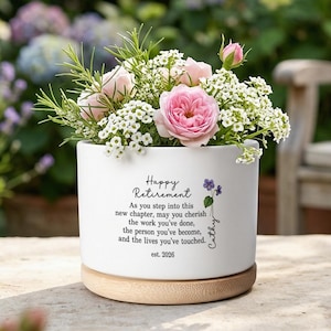 May include: A white ceramic planter with a bamboo base containing pink roses and white flowers. The planter features the text: Happy Retirement, As you step into this new chapter, may you cherish the work you've done, the person you've become, and the lives you've touched, Cathy, est. 2026.