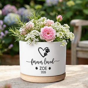 May include: A white cylindrical ceramic planter with a light wood base, filled with pink and white flowers. The front features a black graphic of a heart with paw prints, the text "forever loved", the name "ZOE", and the year "2026".