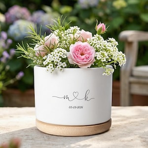 Peut inclure: Un pot de fleurs en céramique blanche avec une base en bois, rempli de roses roses et de fleurs blanches. Le pot porte les initiales "m & k" avec un symbole de cœur et la date "05-09-2026" en écriture noire. Le fond est un jardin.