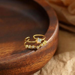 May include: A gold-toned open ring featuring two bands. One band is adorned with a small, iridescent star-shaped accent, while the second band is embellished with a row of tiny, clear, sparkling stones along its length.