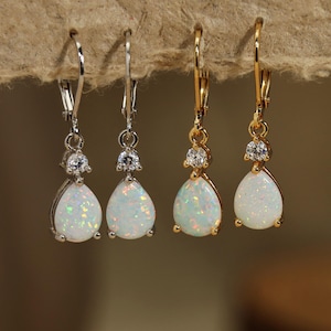 Könnte beinhalten: Vier Paar Tropfenohrringe. Zwei Paare haben silberfarbene Haken und Fassungen, und zwei haben goldfarbene Haken und Fassungen. Jeder Ohrring hat einen kleinen, runden, klaren Stein über einem birnenförmigen, opalisierenden Stein.