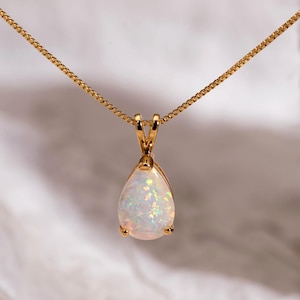Puede incluir: Un collar dorado con un colgante de ópalo en forma de pera. El ópalo tiene una base blanca con destellos iridiscentes de color. La cadena fina cuelga de un pequeño cierre en la parte superior del colgante. El fondo es una superficie suave y blanca texturizada.