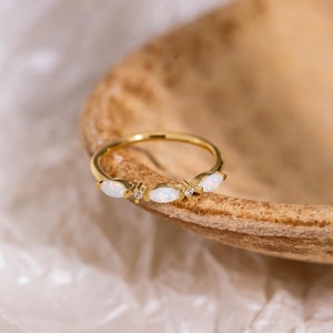 Peut inclure: Une bague en or ornée de trois opales blanches taille marquise disposées horizontalement sur l'anneau, agrémentée de deux petites pierres rondes transparentes placées entre les opales, formant un bijou délicat et symétrique.