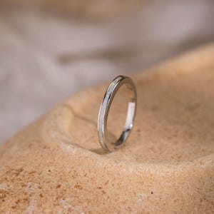 May include: A silver ring with a slender band and a white, iridescent inlay. The ring is displayed against a neutral, textured backdrop, emphasising its delicate design and polished finish. The ring's minimalist style makes it suitable for everyday wear.