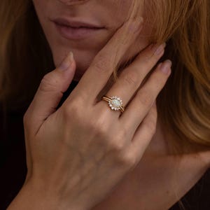 Könnte beinhalten: Ein goldener Ringsatz mit einem ovalen Mittelstein, umgeben von kleineren, funkelnden Steinen. Der Ring befindet sich an einem Finger einer Frau und unterstreicht sein detailliertes Design und seinen eleganten Stil. Der Ring ist ein Schmuckstück.