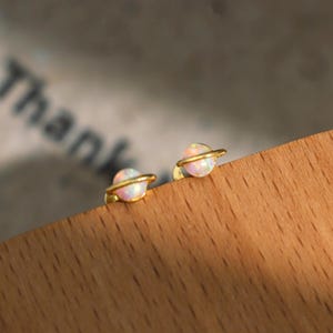 May include: A pair of gold-colored stud earrings, each featuring a small, round, opal-like stone encircled by a thin gold band. The earrings have a planet-like design, with a pearlescent, multi-colored appearance.