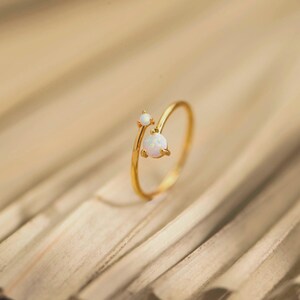 May include: A gold-toned open ring featuring two round, iridescent white opal-like stones of different sizes. The larger stone is set at the end of one band, while the smaller stone is positioned on the other, creating a bypass design.