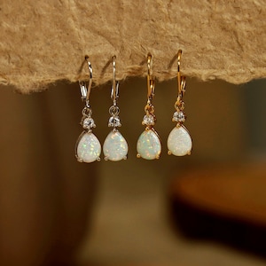 May include: Four pairs of dangle earrings with pear-shaped opal stones. Two pairs have silver-coloured hooks and settings, while the other two have gold-coloured hooks and settings. Each earring features a small, round, clear stone above the opal.