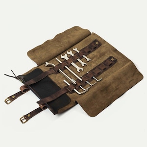 Könnte beinhalten: Eine braune Leder-Werkzeugrolle mit verschiedenen Schraubenschlüsseln und Inbusschlüsseln. Die Rolle besteht aus einem weichen, Wildleder-ähnlichen Material und hat dunkelbraune Lederriemen mit Messingschnallen. Die Werkzeuge sind silberfarben und ordentlich angeordnet.