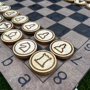 Peut inclure: Un échiquier en bois avec une grille de carrés clairs et foncés, comportant des lettres et des chiffres imprimés en noir sur les bords. Le plateau est garni de pièces de jeu circulaires en bois, chacune gravée d'une icône d'échecs noire.