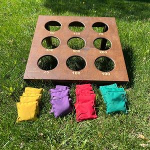 May include: A brown metal toss game board with nine circular holes, each marked with point values 25, 50, 75, 100, 150, 200, 300, and 500. The set includes four stacks of fabric bean bags in yellow, purple, red, and green colours for gameplay.