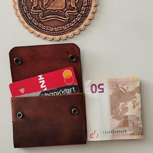Könnte beinhalten: Ein braunes Lederportemonnaie mit zwei Kreditkarten und einem Fünfzig-Euro-Schein. Das Portemonnaie ist geöffnet und zeigt die Karten und das Bargeld. Die Karten sind rot und schwarz, und der Schein ist weiß und beige.