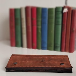 Könnte beinhalten: Eine braune Ledergeldbörse mit zwei Metall-Druckknöpfen. Die Geldbörse ist rechteckig und gefaltet. Im Hintergrund, unscharf, eine Reihe bunter Bücher.