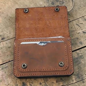 May include: Open, brown leather wallet with a snap closure. The wallet has a card slot and a zippered compartment. The leather has a distressed look, and the wallet is on a wooden surface.