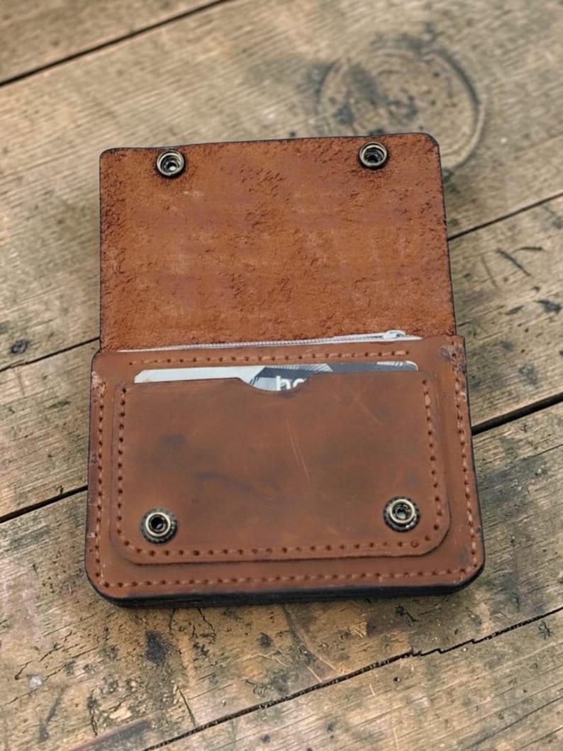 May include: Open, brown leather wallet with a snap closure. The wallet has a card slot and a zippered compartment. The leather has a distressed look, and the wallet is on a wooden surface.