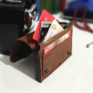 May include: A brown leather wallet with a zipper closure and multiple card slots. The wallet is open, revealing several credit cards and folded bills. The wallet is on a white surface.