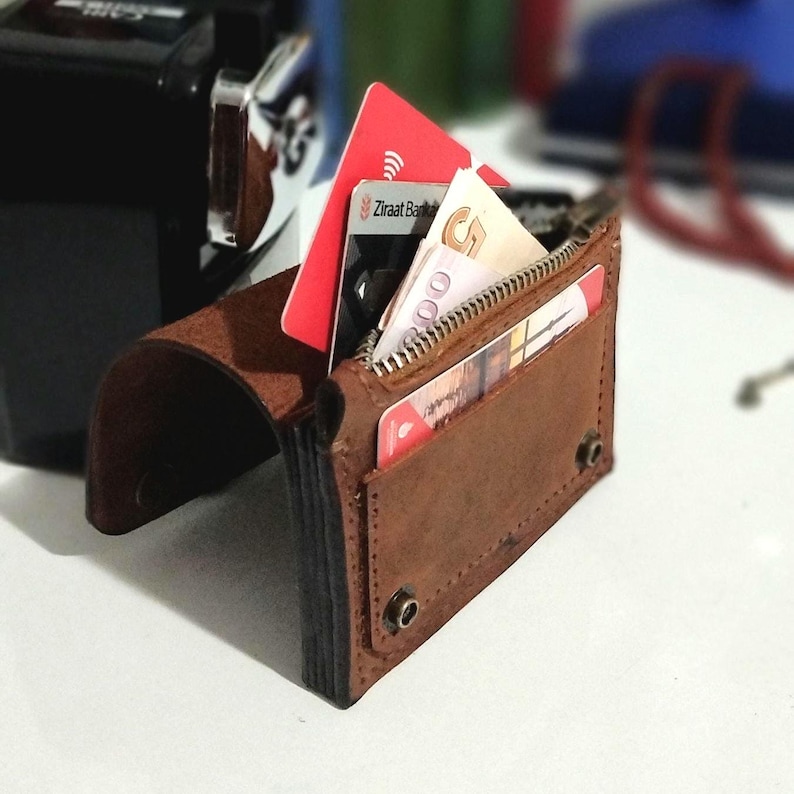 May include: A brown leather wallet with a zipper closure and multiple card slots. The wallet is open, revealing several credit cards and folded bills. The wallet is on a white surface.