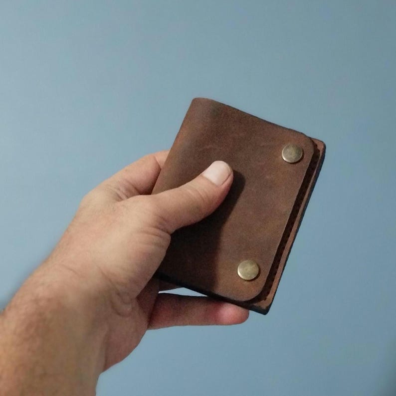 May include: A brown leather wallet with two brass-colored snap closures. The wallet is rectangular and appears to be made of a distressed leather material. The wallet is held in a hand against a light blue background.