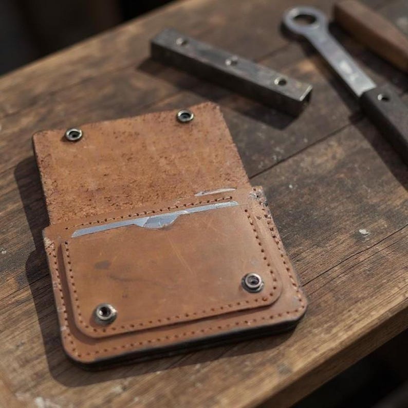 May include: Open, light brown leather wallet with a card slot. The wallet is secured with two metal snaps. The wallet is on a wooden surface, with metal tools in the background.