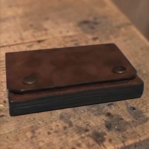 May include: A small, rectangular brown leather wallet with a flap secured by two brass-colored rivets. The wallet appears to be handmade and sits on a wooden surface.