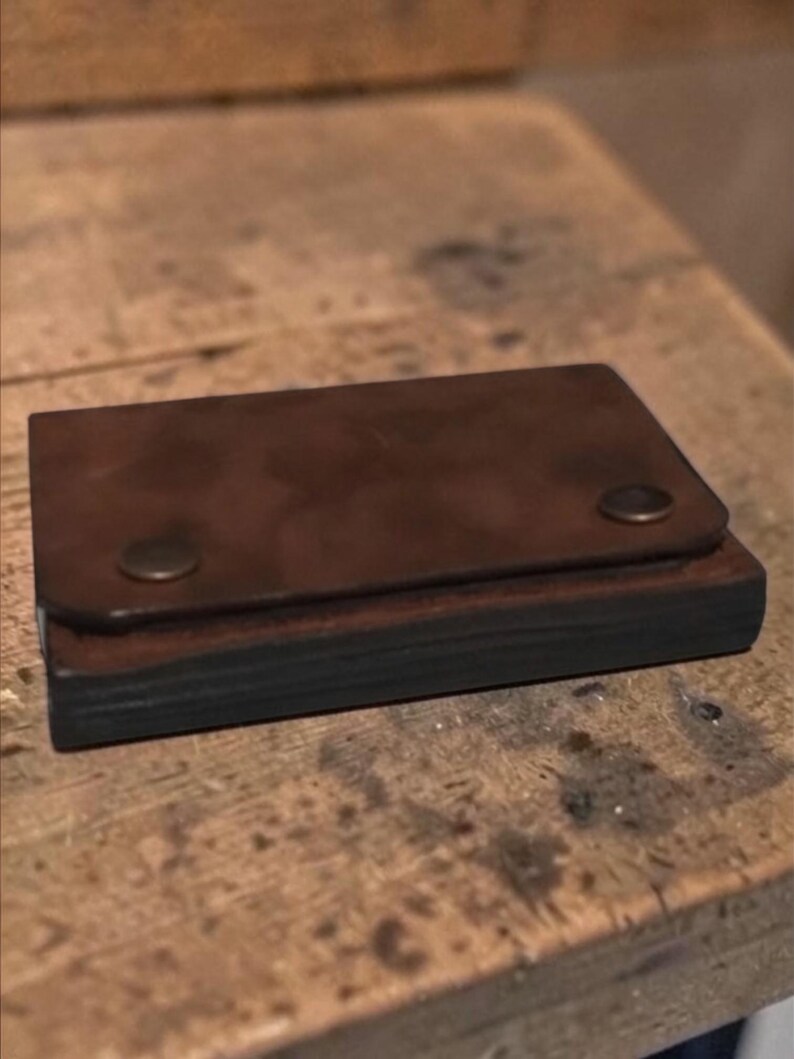 May include: A small, rectangular brown leather wallet with a flap secured by two brass-colored rivets. The wallet appears to be handmade and sits on a wooden surface.