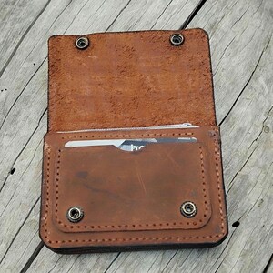 May include: Open brown leather wallet with a snap closure. The wallet has a card slot and a zippered pocket. The leather has a distressed look. The wallet is on a wooden surface.