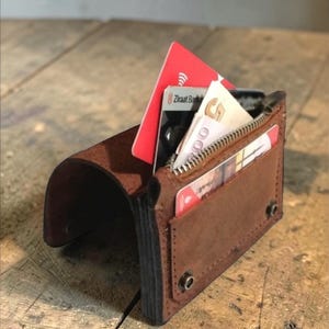 May include: A brown leather wallet with a curved flap and a zippered pocket. The wallet is open, revealing several cards and cash. The wallet is on a wooden surface.