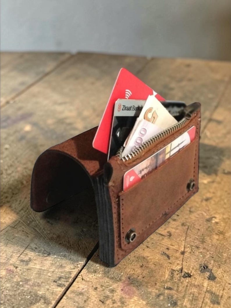 May include: A brown leather wallet with a curved flap and a zippered pocket. The wallet is open, revealing several cards and cash. The wallet is on a wooden surface.
