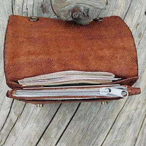 May include: Open, brown leather wallet with a silver zipper and a stack of papers inside. The wallet is open, revealing the interior and contents. The leather has a textured appearance.