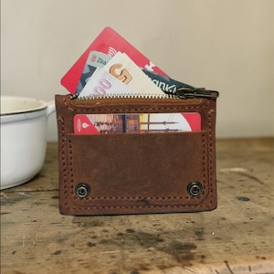 May include: A brown leather wallet with a zipper closure and a front pocket. The wallet is open, revealing a red credit card and Turkish lira banknotes. The wallet is on a wooden surface.