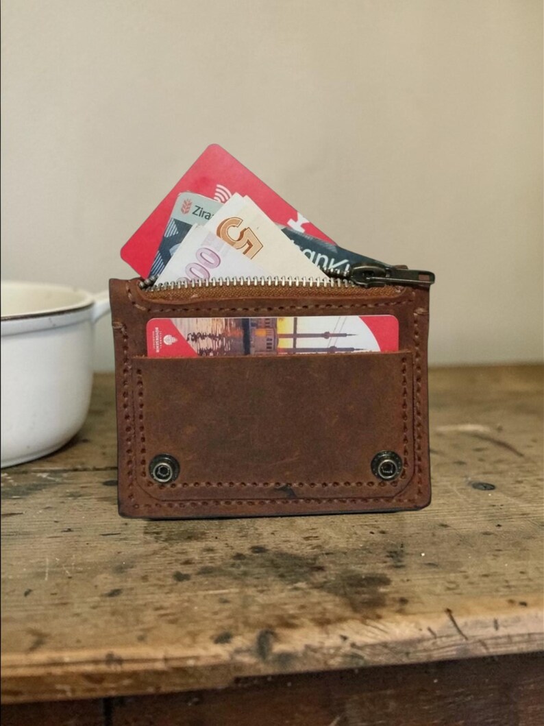 May include: A brown leather wallet with a zipper closure and a front pocket. The wallet is open, revealing a red credit card and Turkish lira banknotes. The wallet is on a wooden surface.
