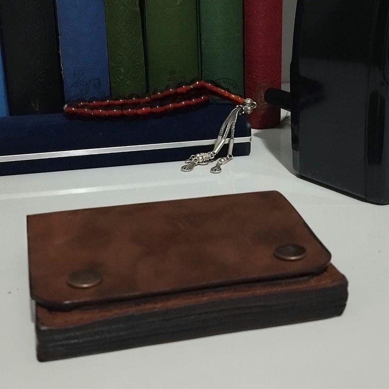 May include: A brown leather wallet with a snap closure. The wallet is rectangular and has a worn appearance. A red beaded strand and a silver ornament are in the background.