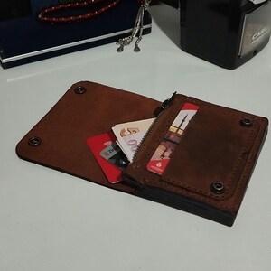 May include: A brown leather wallet with a bi-fold design, featuring a zippered compartment and multiple card slots. The wallet is open, revealing cards and currency. Rivets secure the wallet's construction.