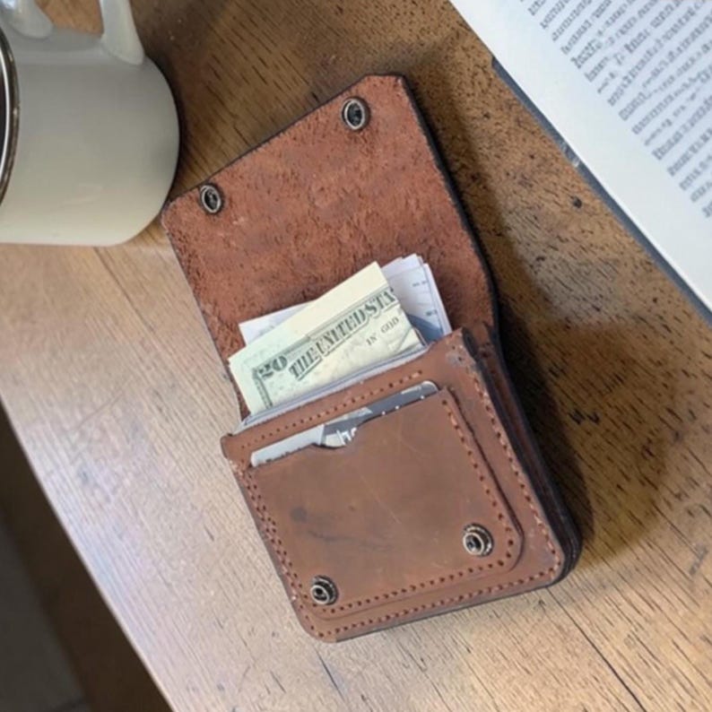 May include: A brown leather wallet with a bi-fold design, secured by snap closures. The wallet is open, revealing a twenty-dollar bill and other contents. The stitching is visible around the edges.