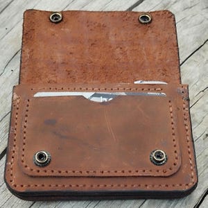 May include: Open brown leather wallet with two snap closures. The wallet has a card slot and is stitched around the edges. The interior is a lighter shade of brown than the exterior.