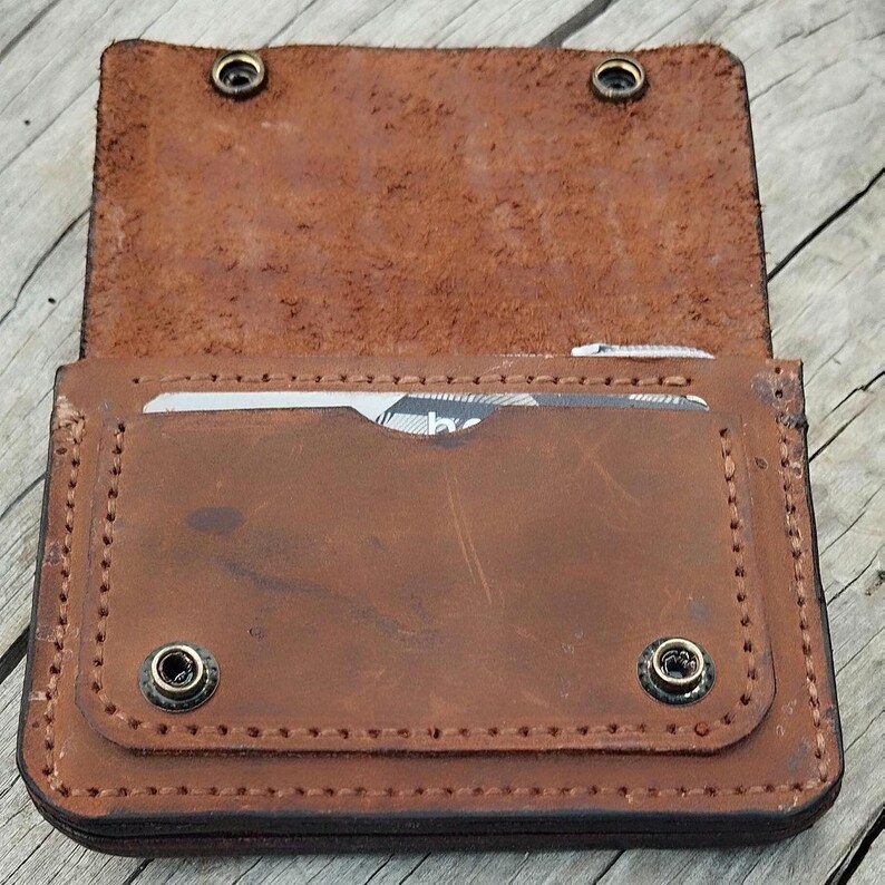 May include: Open brown leather wallet with two snap closures. The wallet has a card slot and is stitched around the edges. The interior is a lighter shade of brown than the exterior.