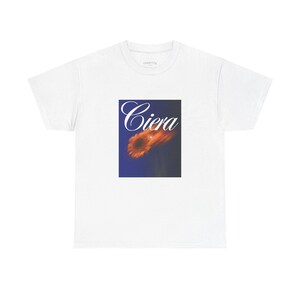 以下が含まれることがあります： 白いTシャツ。筆記体の「Ciera」の文字が、カラフルな抽象的なデザインの上に配置されています。デザインは、濃い青色の背景にオレンジと黄色の色合いが含まれています。カジュアルな普段着のTシャツ。