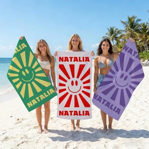 May include: Three beach towels with a smiling sun design and the name "Natalia". One towel is green with a yellow sun, another is white with a red sun, and the third is purple with a lavender sun.
